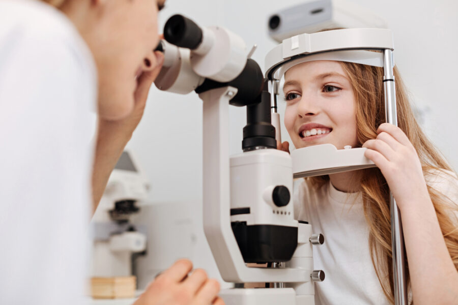 Young girl smiling during a slit lamp eye examination conducted by an optometrist, promoting early detection and pediatric eye health. - Dilated pupils