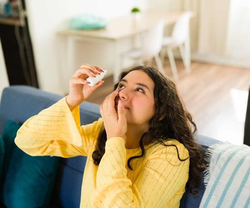 Teenager applying eye drops while sitting on a comfortable sofa in her living room, relieving dry or irritated eyes at home