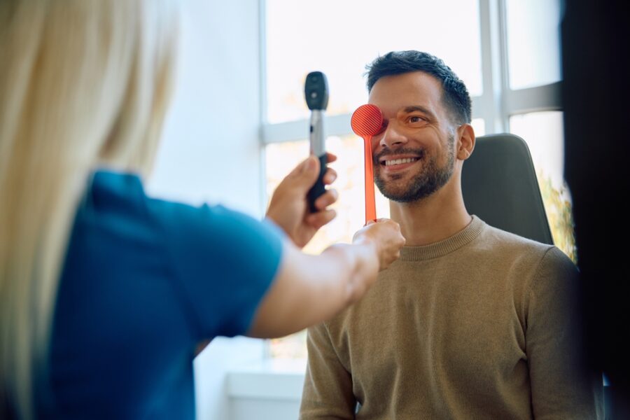 Smiling male patient taking a vision test during an eye exam, with an optometrist using an occluder and ophthalmoscope to check eye health. - Myopia