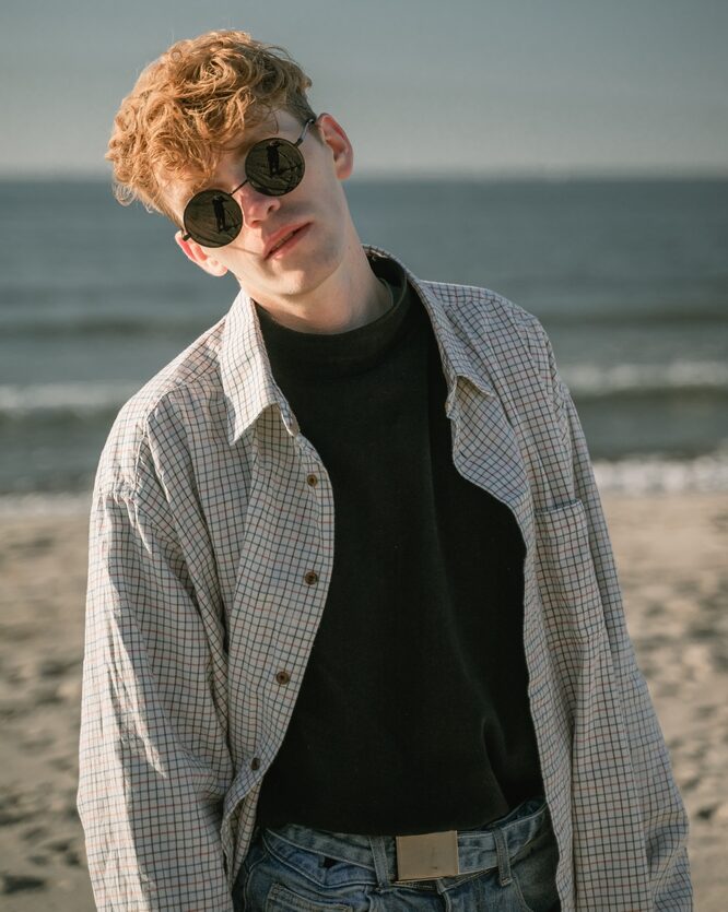 Blonde man standing by the beach wearing checkered polo, black t-shirt for inner and a circular sunglasses - See Breeze Optometry Blonde man standing by the beach wearing checkered polo, black t-shirt for inner and a circular sunglasses - See Breeze Optometry